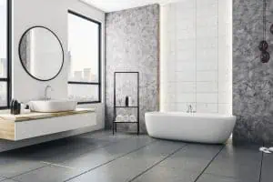 Modern bathroom with a large white freestanding bathtub, a wall-mounted sink with a round mirror, and a view of the cityscape through a window. The interior features gray tiles and walls, designed by an expert painting company in Nassau County NY. A black metal shelf holds towels and decor.