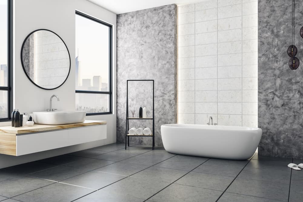 Modern bathroom with a large white freestanding bathtub, a wall-mounted sink with a round mirror, and a view of the cityscape through a window. The interior features gray tiles and walls, designed by an expert painting company in Nassau County NY. A black metal shelf holds towels and decor.