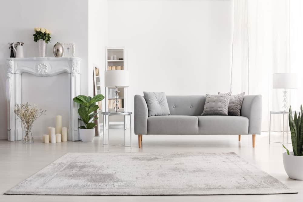 A bright, minimalist living room featuring a gray sofa, decorative pillows, and a white ornate fireplace. Potted plants and candles are artfully placed around the room. Bathed in natural light from large windows, it's the perfect setting for showcasing exquisite interior or exterior painting by a trusted company in Nassau County NY.