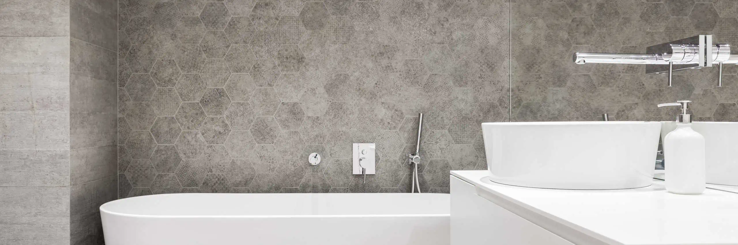 Modern bathroom featuring a white freestanding bathtub, a white sink with a sleek faucet, and a soap dispenser, all set against textured gray hexagonal tiles on the wall. Expertly designed like an Interior Painting Nassau County NY masterpiece, it exudes elegance and contemporary charm.