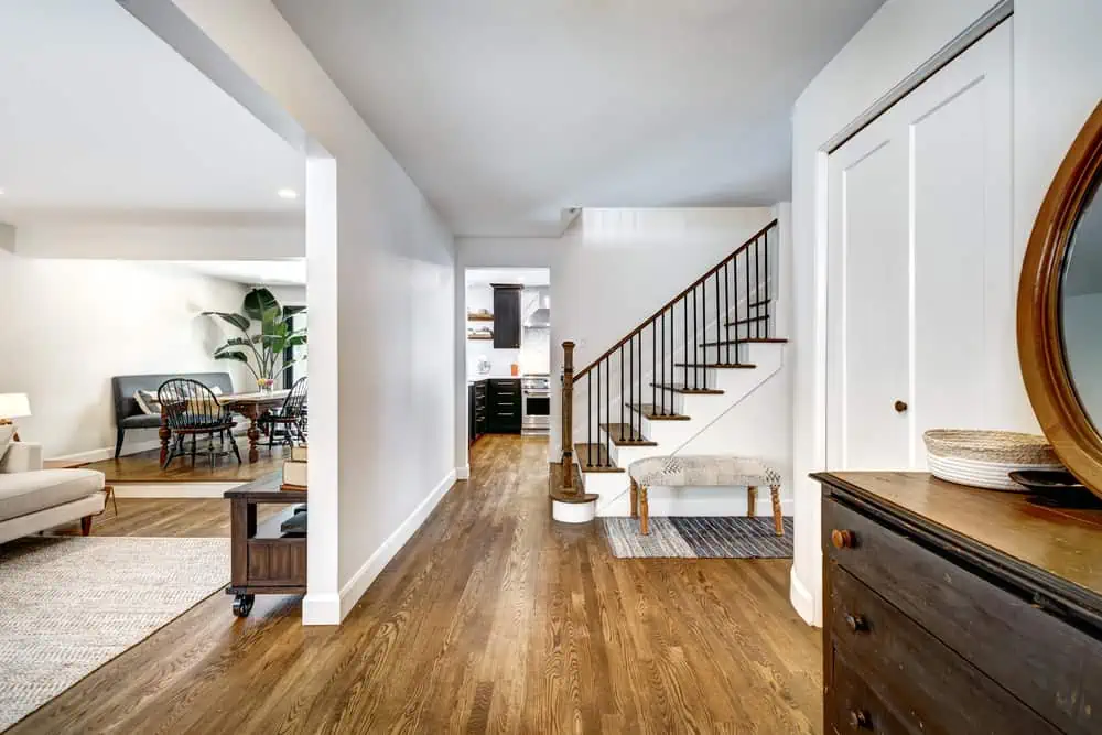 A modern home interior in Nassau County, NY, showcases a hallway with a wooden staircase, bench, and dresser. The open area features a living room with a sofa and dining table. Wooden flooring and neutral-colored walls are complemented by expert residential painting from a local company.