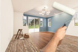 A hand using a paint roller transforms a white-walled room with wooden floors into one with blue walls. The room, set in Nassau County NY, features large windows, a ladder, and a toolbox, with a chandelier hanging from the ceiling. Outside, greenery is visible as the work of an interior painting company unfolds.