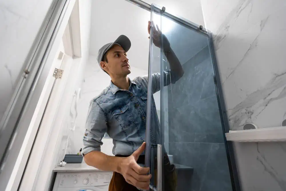 A person wearing a denim shirt and cap is installing or adjusting a glass shower door in a sleek bathroom with marble-patterned walls, perhaps after an interior painting project in Nassau County, NY.