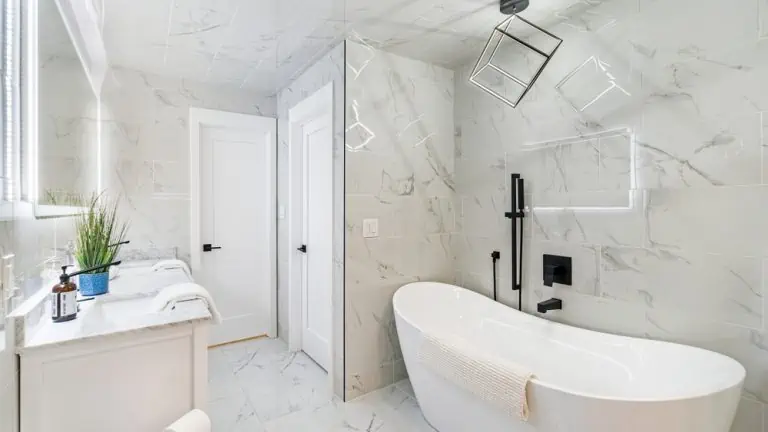 Modern white bathroom with marble tiles features a freestanding bathtub, black fixtures, and a geometric pendant light. A double-sink vanity with white towels and a potted plant under a large mirror is on the left. Perfect for those seeking interior painting in Nassau County, NY, two white doors are in the background.