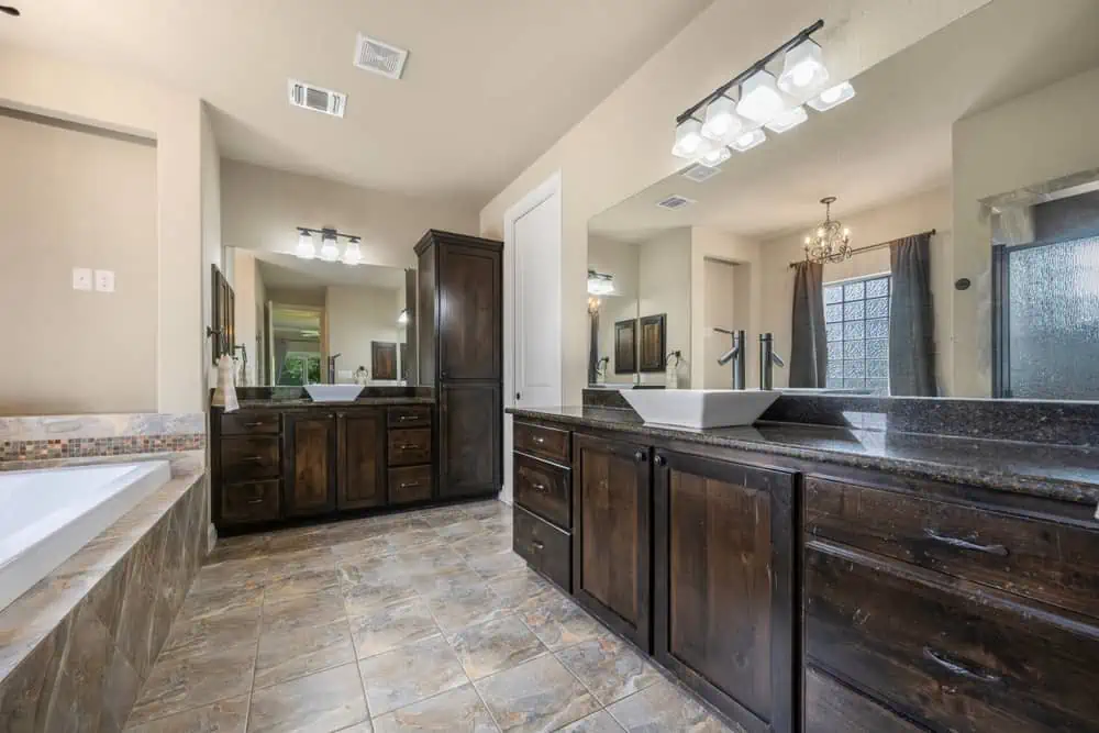 A modern bathroom with dual sinks on a dark wood vanity, a large mirror, tiled flooring, and a built-in bathtub showcases the elegance found in many homes that opt for residential painting in Nassau County NY. Recessed lighting and a chandelier illuminate the space, while a frosted glass shower is visible in the background.