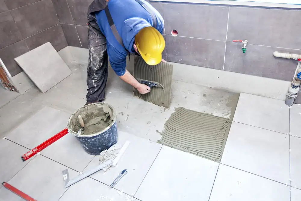 A construction worker in a yellow hard hat and blue sweatshirt is laying gray ceramic tiles on a floor, preparing the elegant base of a home before the Painting Company Nassau County NY arrives. He spreads adhesive with a trowel, surrounded by tools, a bucket, and tiles in the partially tiled room.