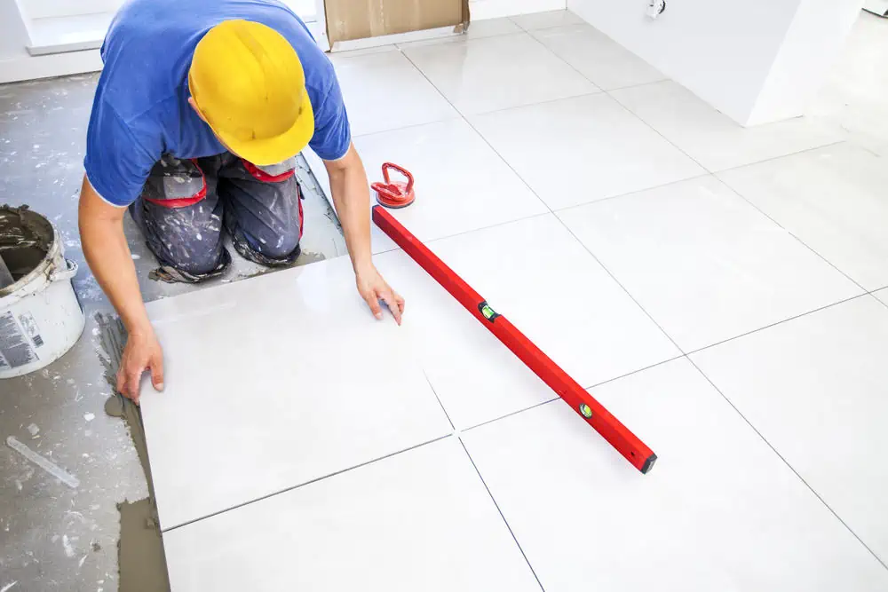 A worker in a yellow hard hat and blue shirt is installing large white floor tiles at an indoor renovation site. A red level tool rests on the tiles, while a bucket of adhesive is nearby, setting the stage for future interior painting in Nassau County, NY.