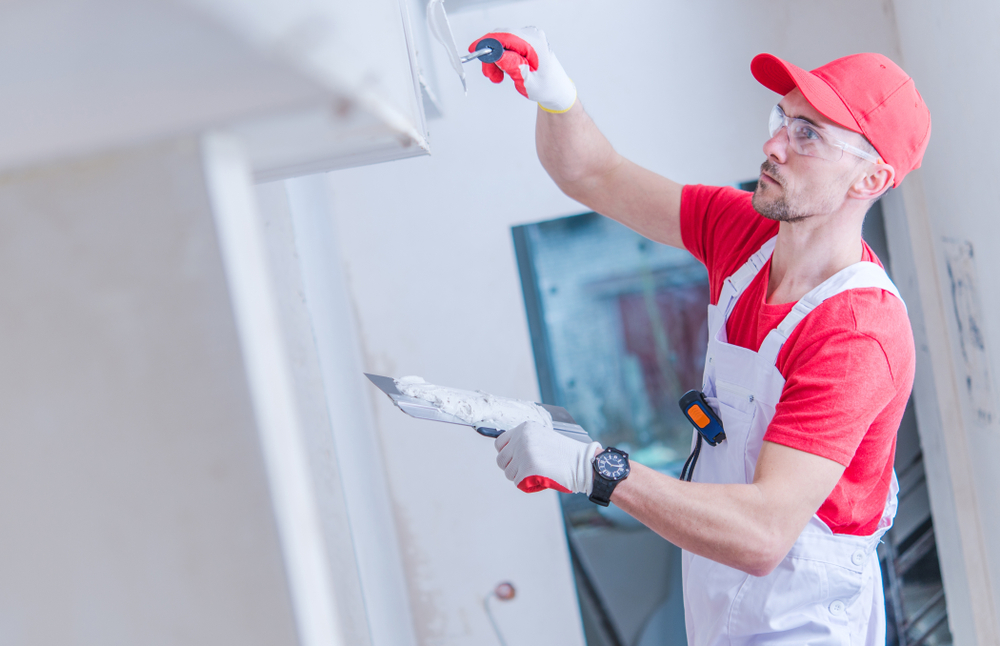 A construction worker in red and white attire applies plaster to a wall using a putty knife. Representing an interior painting company in Nassau County, NY, he dons safety glasses, gloves, and a red cap. The background reveals a partially finished room with a window.
