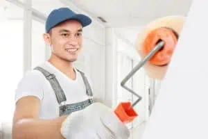 A smiling man in a blue cap and overalls paints a wall with a roller in a bright room, showcasing his expertise in interior painting. He wears white gloves and a t-shirt, framed by large windows that highlight the modern Nassau County NY interior.