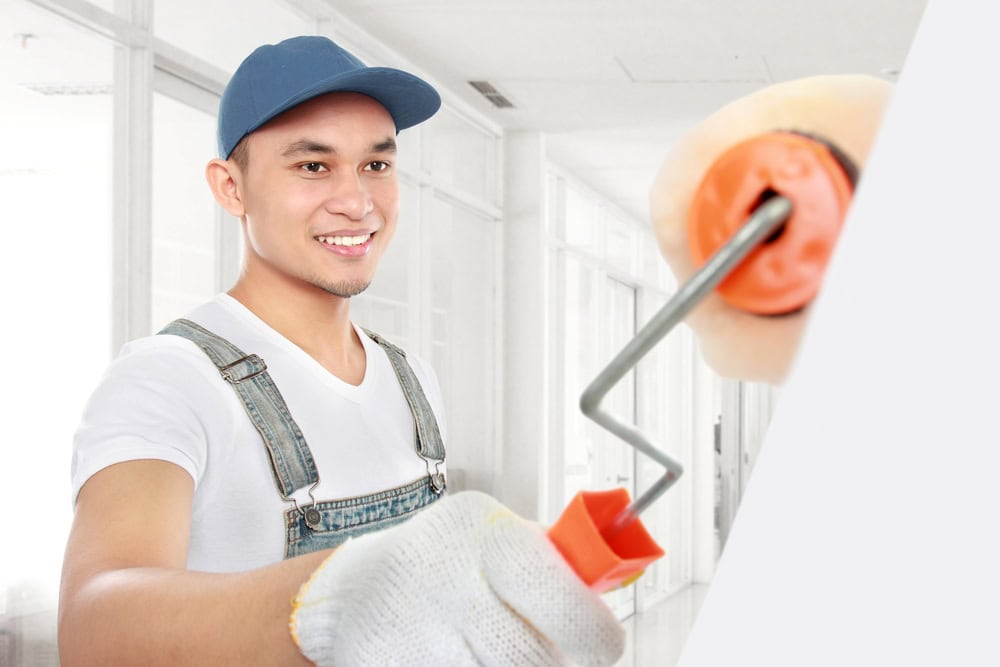 A smiling man in a blue cap and overalls paints a wall with a roller in a bright room, showcasing his expertise in interior painting. He wears white gloves and a t-shirt, framed by large windows that highlight the modern Nassau County NY interior.