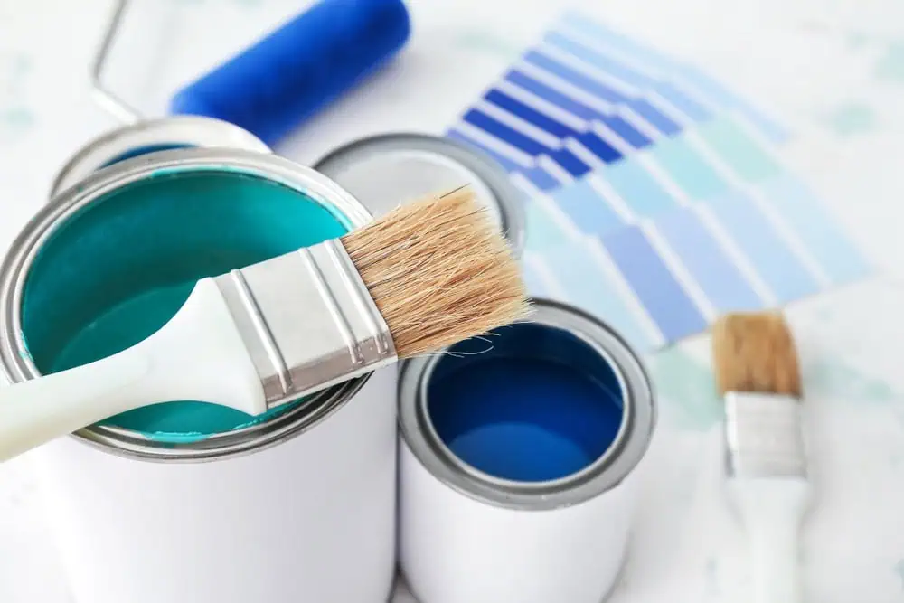 A white paint can with teal paint open, a brush resting on top. Another can of deep blue nearby. A roller sits beside blue swatches, arranged from light to dark—tools set on a light surface, showcasing the art of interior painting in Nassau County, NY.