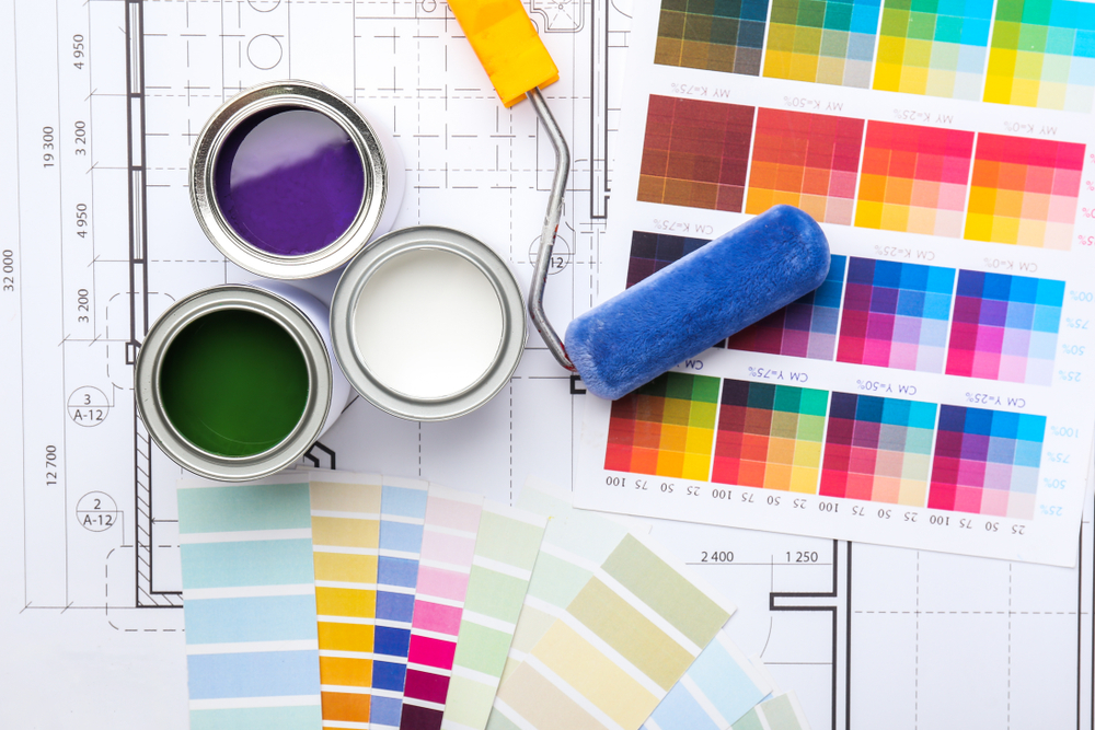 Open paint cans in purple, white, and green with a paint roller rest on architectural blueprints. Various color swatches hint at a design or renovation project underway by a professional residential painting company in Nassau County, NY.