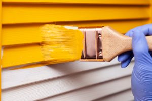 A hand wearing a blue glove is painting a wooden surface with bright yellow paint, embodying the expert touch of a top Painting Company in Nassau County NY. The wooden slats transition beautifully from white to yellow under the skilled brushwork.