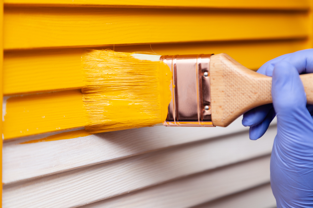 A hand wearing a blue glove is painting a wooden surface with bright yellow paint, embodying the expert touch of a top Painting Company in Nassau County NY. The wooden slats transition beautifully from white to yellow under the skilled brushwork.