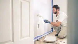 A man in a white t-shirt and beige pants is engaged in interior painting, carefully transforming a wall white using a roller. Kneeling by the paint tray, he has secured the edges with blue tape. A door peeks from the left, witnessing this Nassau County NY transformation.
