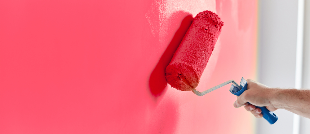 A hand holding a paint roller applies bright red paint to a wall with precision, showcasing the smooth, even layer. This image captures the vibrant artistry of an interior painting process by a skilled Painting Company in Nassau County NY.
