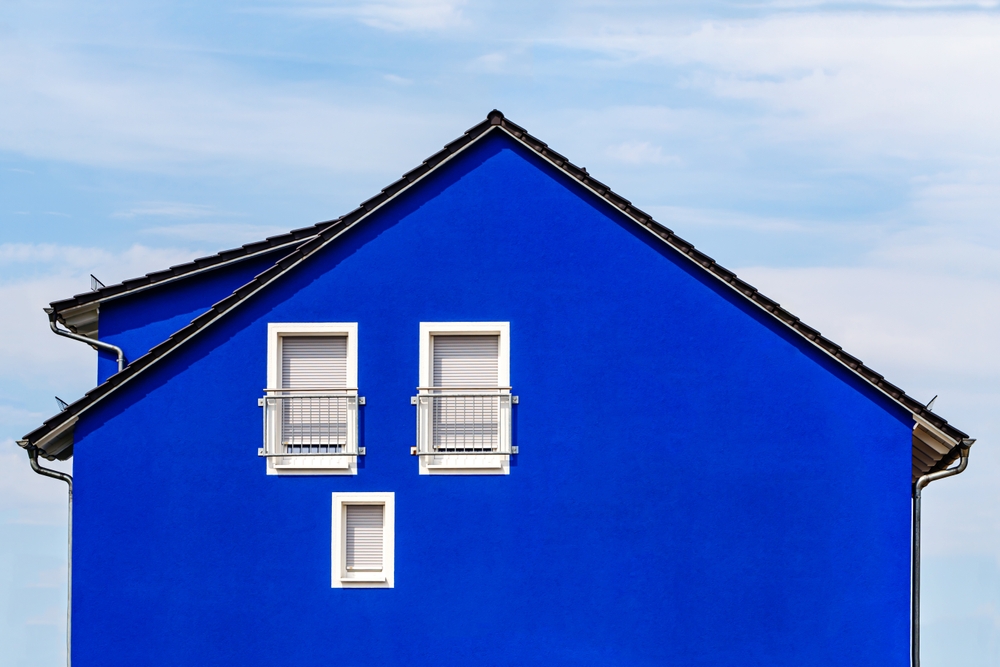 A vibrant blue house facade with a triangular roof stands against a cloudy sky, a testament to the artistry of Residential Painting Commercial Painting Nassau County NY. The house features two small white-framed windows with closed shutters and gray railing on the upper right side.