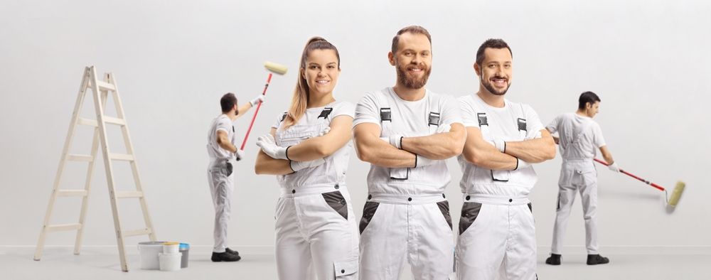 Three painters in white overalls stand with arms crossed, smiling at the camera. Behind them, two people are painting a wall, showcasing their skills in both interior and exterior painting projects for a trusted painting company in Nassau County, NY.