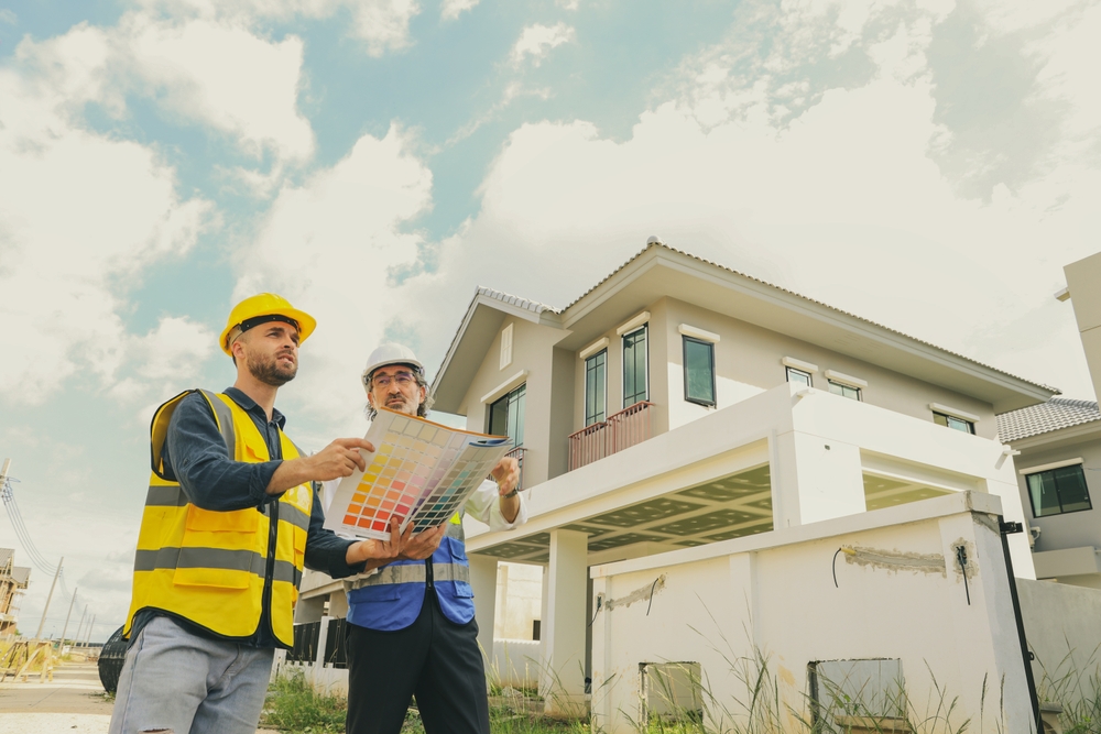 Two construction workers in safety vests and helmets stand outside a newly built house, examining a color chart for an Interior Painting project in Nassau County NY. They discuss plans under a partly cloudy sky. The modern house boasts a light exterior, showcasing the expertise of their Painting Company.