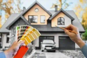 A person holds a paint swatch and brush, carefully selecting colors for the modern home's exterior. Surrounded by blurred autumn foliage, this scene exemplifies residential painting expertise, a hallmark of any reputable painting company in Nassau County NY.