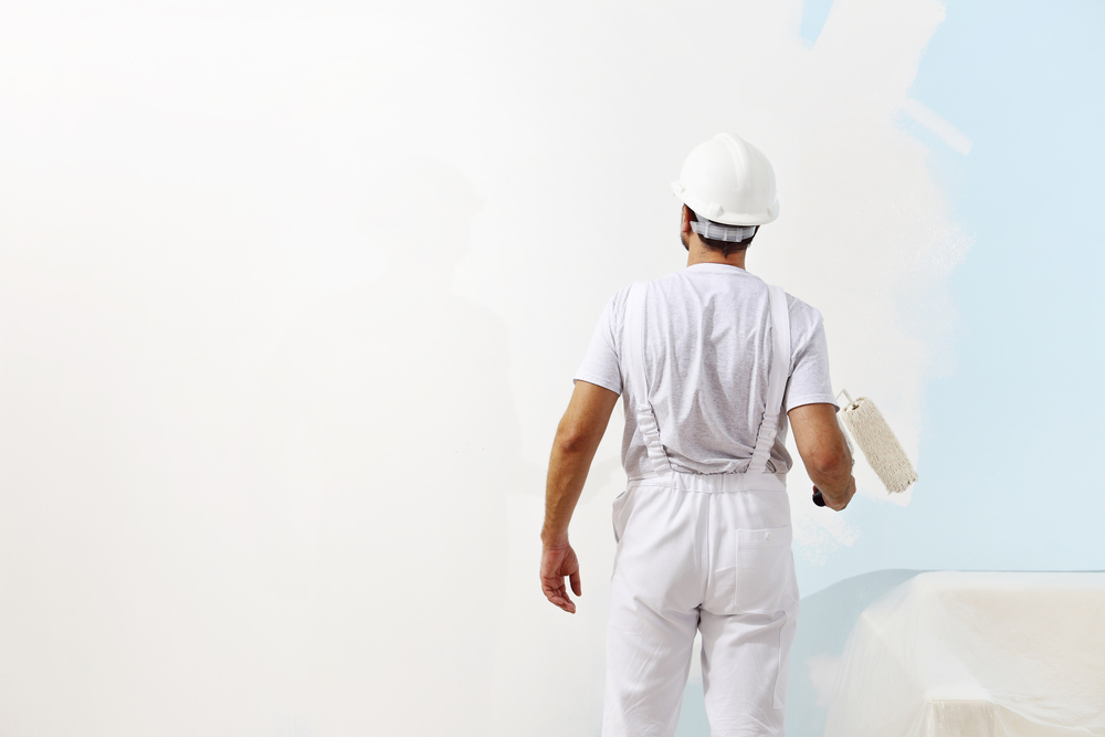 A person wearing white overalls and a hard hat is painting a wall with a roller. The wall transitions from blue to white, and a tarp covers furniture in the lower right corner. This meticulous interior painting project enhances a home in Nassau County, NY.
