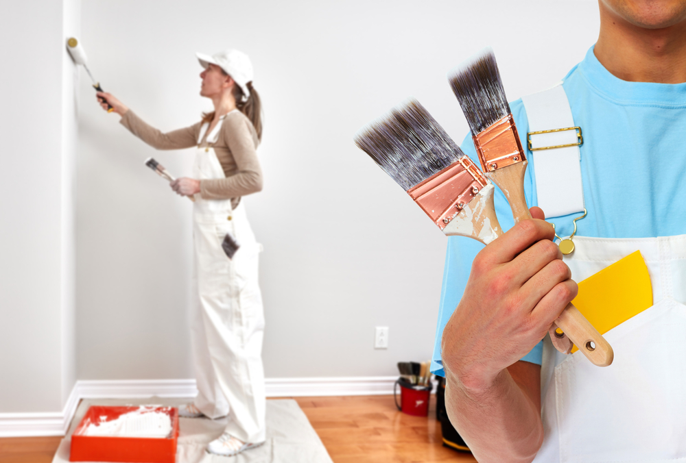 Two painters wearing overalls are working indoors. One is painting a wall with a roller, while the other holds three paintbrushes and a color card. They are surrounded by painting supplies on a wooden floor.