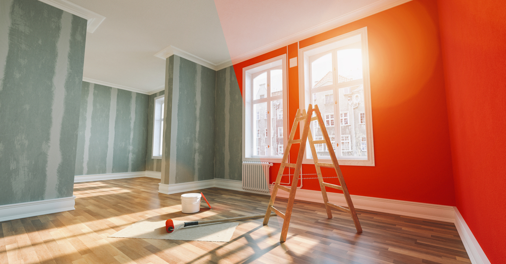 A brightly lit room in Nassau County, NY, with large windows, hardwood floors, and freshly painted red and gray walls. A wooden ladder, paint roller, and paint bucket from an interior painting project are placed on a protective sheet on the floor. Sunlight streams in through the windows.