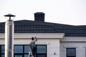 A person on a ladder paints the edge of a modern building's roof, showcasing the skill of a top Painting Company in Nassau County, NY. The building features large windows and a textured black roof with a metallic chimney in the foreground.