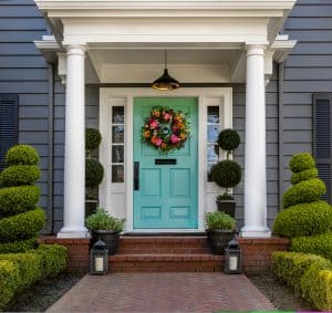 A turquoise front door with a colorful floral wreath is centered between white columns and sidelights. Symmetrical topiary plants and black lanterns frame the brick porch, with manicured hedges lining the entryway.